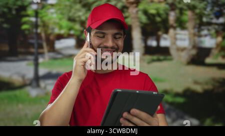 Man wearing red cap smiling while holding smartphone to ear and reading tablet on street; connection. Stock Photo
