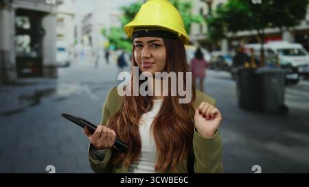 Hispanic woman architect wearing a hardhat stands outdoors on a bustling street, holding a tablet and gesturing, embodying confidence and professional Stock Photo
