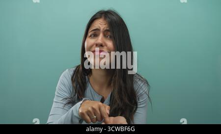 Young hispanic woman beckons open palm for money in studio against green wall, making beckoning gesture; urgent plea. Stock Photo