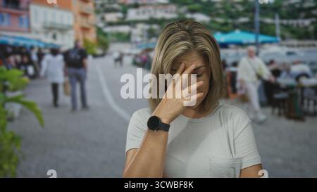 Young blonde woman wearing smartwatch and casual tee pinches nose with hand amid passersby on street; disgust. Stock Photo