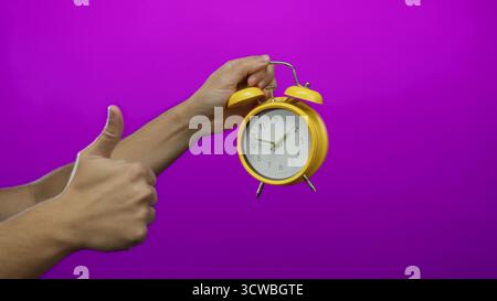 Man holding yellow clock against a vibrant purple wall, with arm pointing, creating a focused and colorful isolated composition. Stock Photo