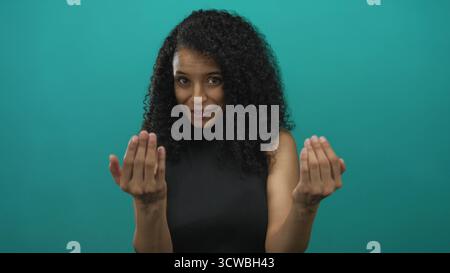 Young hispanic woman with open palms beckons in studio against teal wall with warm smile; friendly invitation. Stock Photo