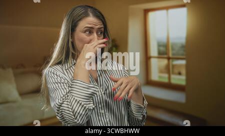 Middle aged blonde woman pinches nose beside window in spacious yellow building lobby wearing striped shirt; disgust. Stock Photo
