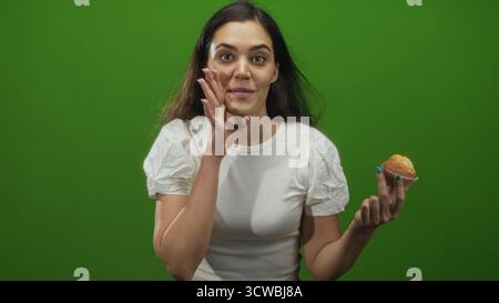 Young hispanic woman holding muffin beckons with open hand in green studio, smiling broadly and speaking to camera; playful invitation. Stock Photo