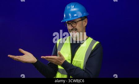 Man in hardhat and safety vest gesturing confidently against a blue background, depicting skilled professional engaging in a presentation or instructi Stock Photo
