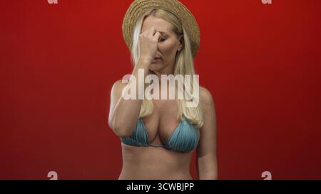 Woman with blonde hair wearing straw hat and blue bikini top pinches nose on red studio set; disgust. Stock Photo