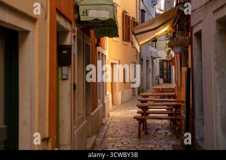 Rovinj, Croatia - August 15, 2025: A narrow cobblestone alley in Rovinj lined with colorful facades, empty wooden café tables, and warm evening sunlig Stock Photo