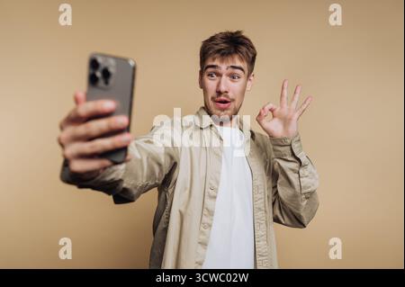 portrait of young surprised man captures a selfie with his smartphone, making an 'okay' gesture on beige background Stock Photo