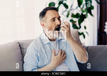 Mature man using an inhaler for breathing support at home while sitting on a sofa with a calming indoor atmosphere around Stock Photo