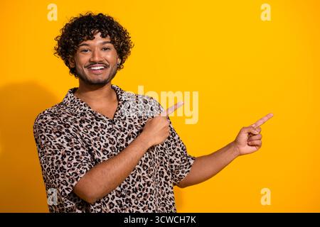 Young man in a leopard print shirt smiles and points against a vibrant yellow background, conveying joy and stylish energy Stock Photo