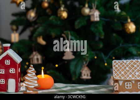 Gingerbread tree cookie with zigzag icing beside orange candle, red ceramic house and gingerbread house on table with Christmas tree. Concept of mixed Stock Photo
