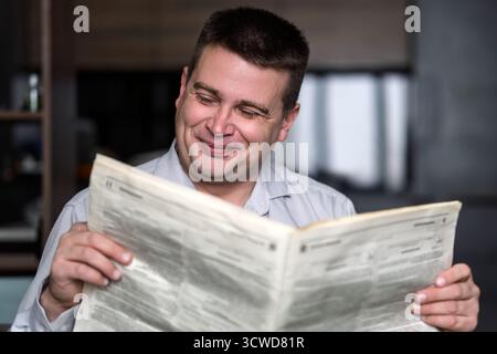 Smiling mature man reading newspaper while sitting at the table on a dark background. Stock Photo