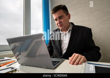 A young handsome man in black suit is working at a computer on a light background. the nerd looks at the monitor screen Stock Photo