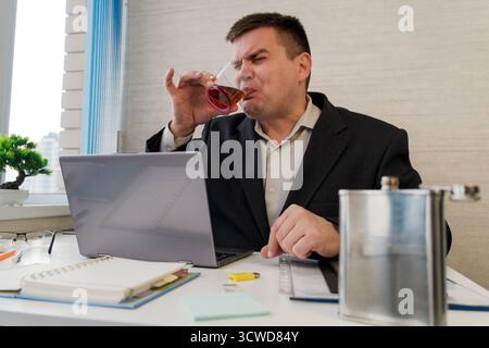 A businessman drinks alcohol while sitting at a table next to a laptop in a office. Drink alcohol in the workplace. Alcoholic employee. Stock Photo