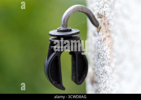 A close-up shot of a black plastic insulator with a curved metal hook attached to a textured, light-colored surface, with a blurred green background. Stock Photo