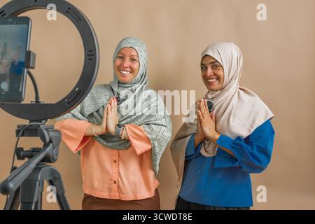 Two smiling women wearing hijabs doing namaste gesture in front of ring light and phone Stock Photo