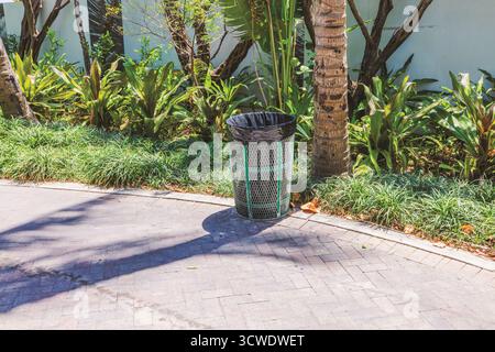 Metal trash bin with black bag near palm trees and green plants on Ocean Drive in Miami Beach. USA. Stock Photo