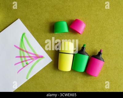 Closeup of highlighter pens of different bright colors and white paper with fluorescent pink and green marker lines on green cloth. Flatlay with copy Stock Photo