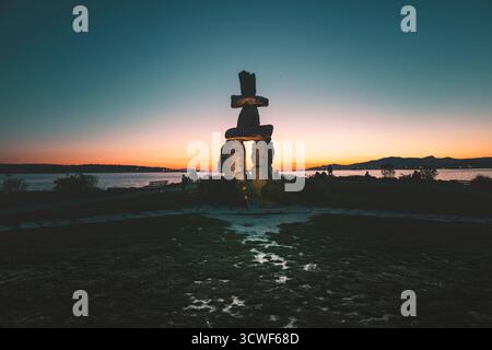 A beautiful sunset sets the sky aglow behind a large stone sculpture near the waterfront. The scene captures peaceful nature at dusk. Stock Photo