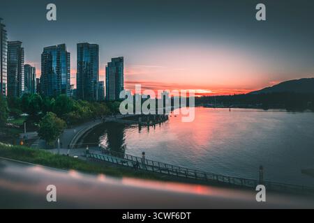 The vibrant sunset creates a colorful reflection on the calm water in a bustling city, framed by tall buildings and lush parks. Stock Photo