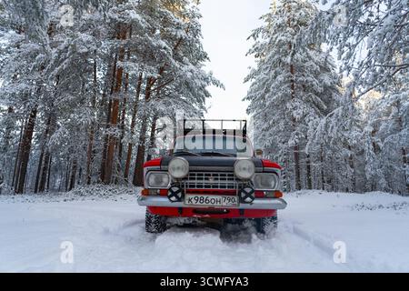 Murmansk Oblast, Russia - December 13, 2024: Low angle frontal view of a classic red Moskvich station wagon rally car in deep snow, framed by tall, sn Stock Photo