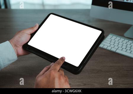 Hands holding a digital tablet with blank white screen on wooden desk, finger touching display, workspace with keyboard in background, copy space idea Stock Photo