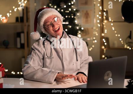 Concept of telemedicine, telehealth, medicine and holidays. Young female doctor in Santa hat and eyeglasses working on laptop computer smiling with po Stock Photo