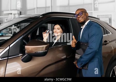 Happy woman in professional attire holds car keys inside vehicle, while man in suit gives thumbs up, representing a positive car buying experience and Stock Photo