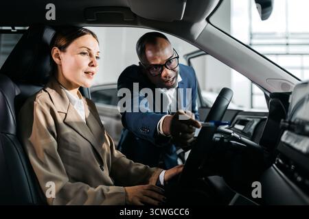 Female professional in beige suit interacts with male colleague, exploring car features and technology in a contemporary automotive setting, highlight Stock Photo