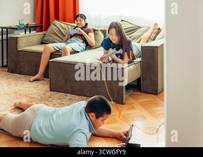 boy on floor playing with tablet while two girls relax on sofa using smartphones with orange curtains and daylight, technology education leisure Stock Photo