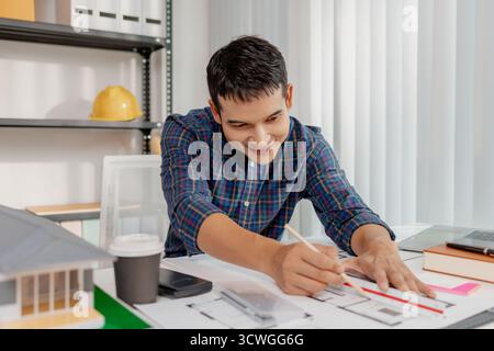 A young male architect is working with blueprints at his desk, using rulers and drawing tools to refine details. He appears focused and engaged, balan Stock Photo