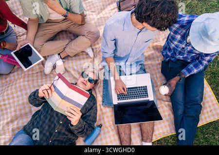 High angle view of a young diverse group of students working on a project with a laptop and books while relaxing on a picnic blanket outdoors. Stock Photo