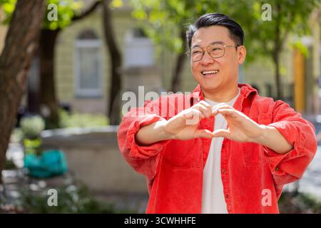 I love you. Asian middle-aged man makes symbol of love, showing heart sign to camera express romantic feelings, express sincere positive feelings outdoors. Charity, gratitude, donation. on city street Stock Photo