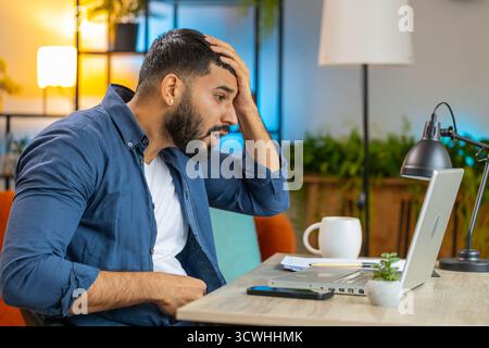 Angry furious Hispanic man freelancer using laptop while working at home office having nervous breakdown at work, stress management, mental distress problems, losing temper, reaction on failure. Stock Photo
