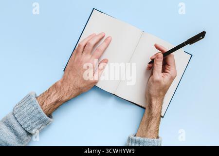Top view of hands writing (Mockup) with a black pen in an open notebook with blank pages. Blue background. Journaling, creative writing, notes, Stock Photo