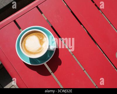 A cappuccino in a turquoise ceramic cup and saucer sits on a vivid red slatted wooden outdoor cafe table in bright sunlight. The coffee features simp Stock Photo