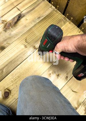 Person using a Bosch cordless drill to secure wooden planks on a deck, focusing on hand and tool against the textured wood surface Stock Photo