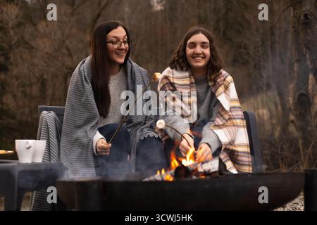 Two young women sitting near a campfire, holding sticks with marshmallows, roasting them and laughing Stock Photo