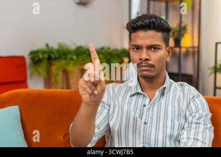 Indian man at home waves finger no no to camera and shows refusal and disagreement with stern mood. Arabian guy on sofa shakes head denies request points finger and shows unhappy sad reject reaction Stock Photo