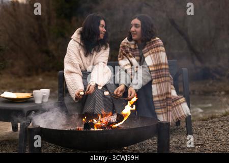 Two women wrapped in blankets sitting outside by the campfire, roasting marshmallows on sticks. Stock Photo