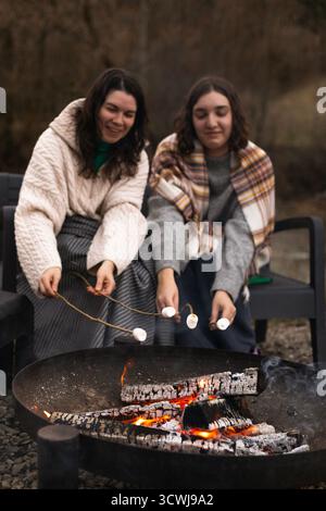 Two women smile thoughtfully while sitting in front of a campfire, roasting marshmallows Stock Photo