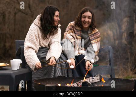 two young women laughing while roasting marshmallows over a campfire Stock Photo