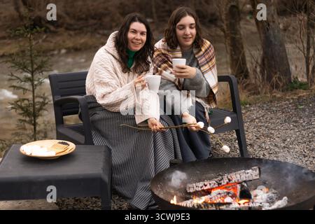 two women by a campfire roasting marshmallows and drinking cocoa Stock Photo