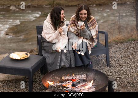 two women wrapped in a blanket sitting by a campfire, laughing, drinking cocoa and roasting marshmallows Stock Photo