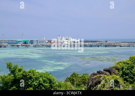 Working piers at Gimnyeong Port line a turquoise basin beside a green‑roof shed and service buildings, with fishing boats tied alongside and breakwate Stock Photo