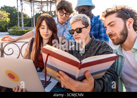 Young diverse group of university students collaborating on a project with a laptop and notebook while sitting together in a city park. Stock Photo