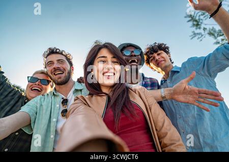 Happy group of young multiracial friends laughing while taking a fun selfie with a smartphone in a park at sunset. Stock Photo