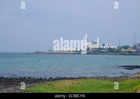 A red lighthouse marks the harbor entrance near an industrial plant and storage silos on Jeju’s northeast coast, viewed across blue‑green water and da Stock Photo