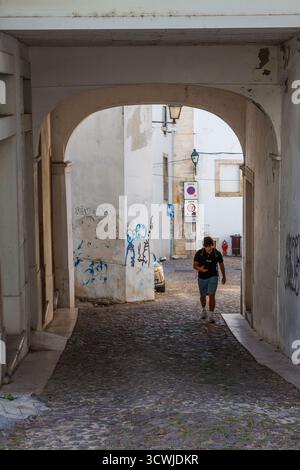 A young man walks through a sunlit, graffiti-marked stone archway onto a cobblestone street in a historic portuguese town, framed by weathered walls a Stock Photo