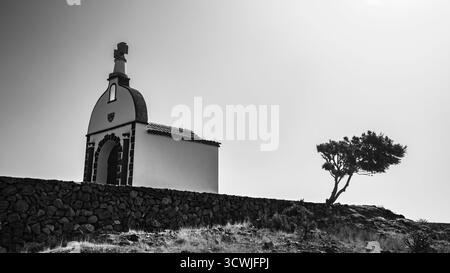 The serene Chapel of San Isidro stands alone atop a hill, framed by a solitary tree in soft light, Alajero, La Gomera, Spain Stock Photo